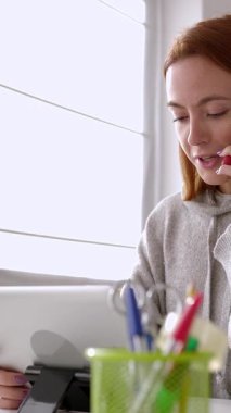 Dijital tablet kullanırken cep telefonuyla konuşan genç bir kadının evdeki ofiste otururken çekilmiş dikey HD videosu. Küçük işletme ve teknoloji yaşam tarzı kavramı.