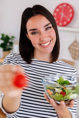 Modern bir mutfakta sağlıklı karışık salata yiyen ve besleyici yemeğin tadını çıkaran genç ve mutlu bir kadının dikey çekimi. Sağlıklı beslenme alışkanlıkları yaşam tarzı ve yiyecek konseptini sağlar.