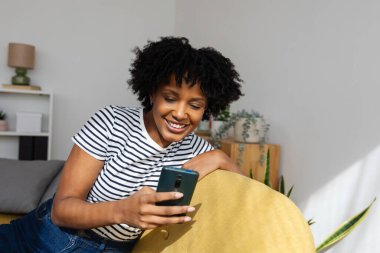 Evdeki rahat bir koltukta akıllı cep telefonunu kullanarak gülümseyen siyah bir kadın sosyal medyada geziniyor ve doğal ışıkla dolu sıradan bir oturma odasında diğerleriyle iletişim kuruyor.
