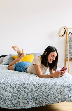 Yatakta cep telefonu kullanan mutlu genç bir kadının dikey görüntüsü. Rahatlamış güzel milenyum dişisi cep telefonuyla sohbet ediyor evde dinleniyor.