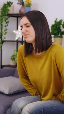 Genç ve güzel bir kadının evdeki koltukta oturup karın ağrısı çektiği gözlerini kapadığı ve karnına sarıldığı dikey HD video. Regl, mide ağrısı, adet konsepti.