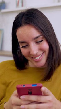 Kanepede oturan mutlu genç kadının dikey HD videosu. Milenyum kadını evde dinleniyor cep telefonundan sosyal medya içeriğini kontrol ediyor. Teknoloji ve eğlence kavramı