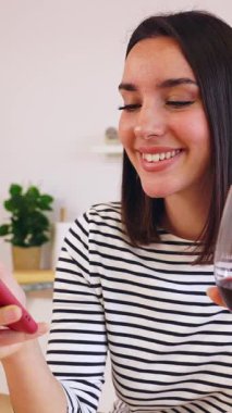 Modern mutfaktaki akıllı telefonu kullanarak evde yemeğinin pişirilmesini beklerken kırmızı şarap içen genç ve mutlu kadının dikey HD videosu.