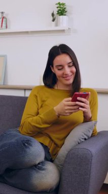 Kafkasyalı mutlu bir kadının cep telefonu kullanarak koltukta dinlendiği dikey HD video. Milenyum kadını elinde cep telefonu cihazıyla kanepede dinleniyor, sosyal medya ağlarından içeriği izliyor.