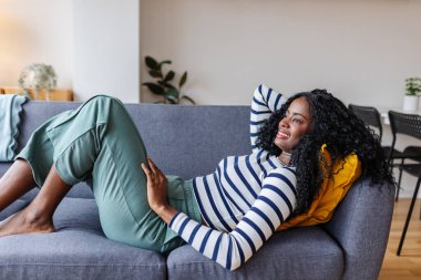 Serene genç siyah kadın rahat bir kanepeye uzanıp evde tatilin tadını çıkarıyor..