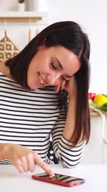 Neşeli, güzel bir kadının akıllı cep telefonunu kullanarak evdeki rahat bir mutfakta oturduğu ve sabahları sosyal medyanın keyfini çıkardığı dikey HD videosu.