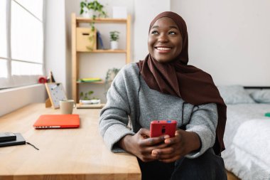 Evde cep telefonu kullanan, başörtüsü takan neşeli, siyah bir kadın. Sosyal medya, gençlik yaşam tarzı ve teknoloji kavramı.