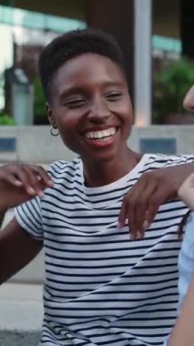 Açık havada oturup gülen ve konuşan neşeli çoklu etnik gençlerin dikey HD videosu. Genç çok ırklı öğrenciler birlikte eğleniyorlar.