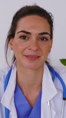 Masa başında oturan genç bayan doktorun gülümseyen portresinin dikey HD videosu. Kadın çevrimiçi danışmanlık hastalarının bakış açısı. Web kamerası veya laptop ekran görüntüsü 
