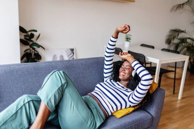 Genç mutlu siyah kadın dans ediyor ve rahat bir kanepede uzanmış evde tatilin tadını çıkarırken çevrimiçi müzik dinliyor..