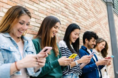 Bir grup farklı arkadaş cep telefonu kullanıyor ve gülümsüyor, şehir ortamında bir tuğla duvarın yanında duruyorlar. Gençlik ve teknoloji yaşam tarzı konsepti