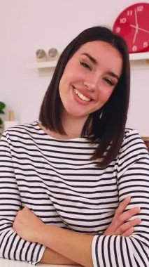 Mutfak masasında oturan ve kameraya gülümseyen mutlu genç kadının dikey HD video portresi.