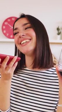 Neşeli, genç ve güzel bir kadın, modern mutfakta kırmızı şarap içerken arkadaşına telefonla fotoğraf ve sesli mesaj gönderiyor. Teknoloji ve ev hayatı kavramı. Dikey HD video