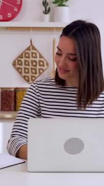 Genç yetişkin bir kadının mutfakta dizüstü bilgisayar kullanırken not aldığı dikey HD videolar. Milenyum kadınlarının çevrimiçi sanal video dinlemeyi öğrenmesi. İş ve eğitim kavramı.