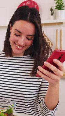 Genç bir kadının sağlıklı bir salatadan hoşlandığı ve modern mutfağında arkadaşlarıyla çevrimiçi ilişki kurduğu dikey HD video.