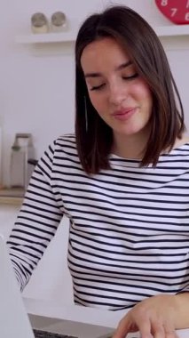 Modern mutfakta uzaktan bilgisayarla çalışan mutlu genç bir kadının sabahları kahve ve akıllı telefonlu dikey HD videosu.