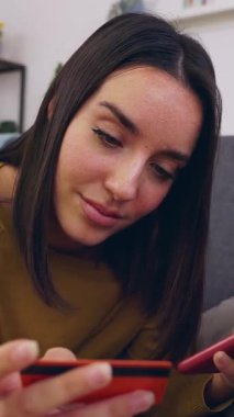 Kanepede uzanan telefon uygulaması üzerinden internetten ödeme yapan genç bir kadının dikey HD videosu. Milenyum güzeli kadın internetten alışveriş yapıyor evden uzaktan ödeme yapıyor..