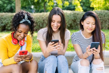 Üç neşeli bayan arkadaş parkta oturmuş, akıllı telefonlarını kullanarak bir yaz gününün tadını çıkarıyorlar.