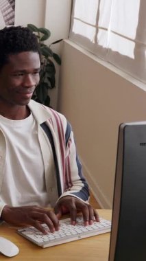 Genç siyahi profesyonelin uzaktan kablosuz kulaklıkla çalışıp, bilgisayarında parlak bir şekilde daktilo yazarak doğal ışık ve bitkilerle organize ettiği dikey HD videosu.