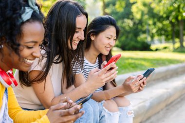 Üç mutlu çoklu etnik kız öğrenci, güzel bir yaz gününde parkta merdivenlerde otururken akıllı telefonlarını kullanıyorlar.