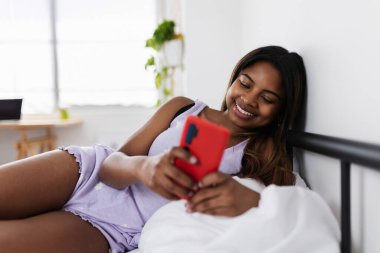 Pijamalı mutlu genç bir kadın, rahat bir şekilde yatakta uzanıyor, sakin bir sabah atmosferinde evde dinlenirken akıllı telefonunun keyfini çıkarıyor.