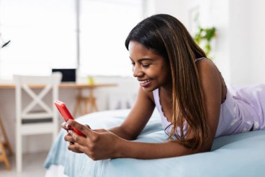 Genç Afro-Amerikan öğrenci kız evde yatakta dinlenirken akıllı telefon kullanıyor.