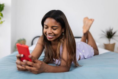 Gülümseyen genç bir kadın cep telefonu kullanarak yatakta uzanıyor, online iletişimden hoşlanıyor ya da evde sosyal medyada geziniyor.