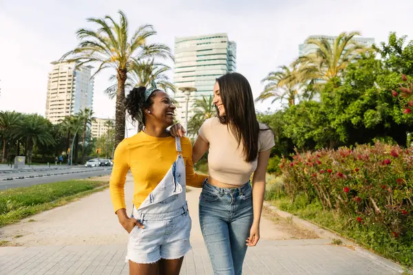 İki genç kadın, hareketli bir şehir parkında, yazın sıcacık parıltısıyla yıkanmış, kol kola yürürken kahkahalar ve sohbetlerini paylaşıyorlar.