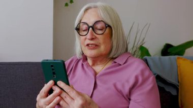 Gülümseyen yaşlı kadın evdeki rahat koltukta otururken cep telefonu kullanıyor, modern teknoloji ve iletişimin tadını çıkarıyor. Büyükannemin cep telefonundan bir video görüşmesi var. Sosyal medya kavramı 