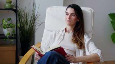 Orta yaşlı bir kadın, rahat beyaz bir sandalyede oturmuş, evinde huzurlu bir rahatlama ve sakin bir zaman geçirirken kitap okuyor.