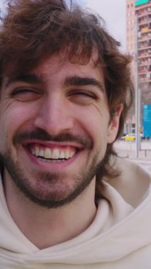 Neşeli genç bir adamın neşe saçtığı dikey HD videosu samimi bir şekilde gülerken, şehir binaları ve arka planda sokak manzaralarıyla canlı şehir manzarasına karşı duruyor.