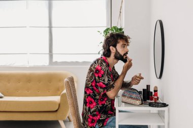 Modern bir dairede dudak kremi süren şık bir genç adam. Dövmeleri özgüven ve orijinallik gösteriyor. Sabah rutini için parlak, havadar bir atmosfer. LGBTQ yaşam tarzı kavramı