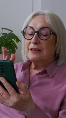 Tekerlekli sandalyedeki neşeli yaşlı kadının ailesiyle görüntülü konuşma yapmak için akıllı telefonu kullandığı ve evdeki modern iletişimin keyfini çıkardığı dikey HD videosu. İletişim yaşam tarzı kavramı 