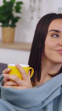 Evdeki kanepede oturmuş, kış günü pencereden dışarı bakarken sıcak kahve içen genç bir kadının dikey HD videosu. Güzel kız sıcak çayın keyfini çıkarıyor kanepede dinleniyor