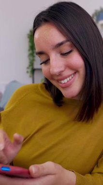 Genç mutlu bir kadının cep telefonundan sosyal medya içeriğini izlerken eğlendiği dikey HD video. Güzel bir milenyum kadını cep telefonu uygulamasında sohbet ediyor evdeki kanepede dinleniyor.