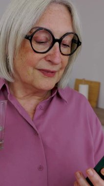 Modern mutfağında akıllı telefon cihazı kullanırken gözlüklü yaşlı bir kadının bir bardak soğuk su içtiği dikey HD video. 