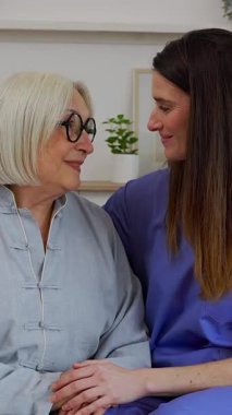 Genç ve mutlu bir kadın hemşirenin evdeki bir kanepede rahat bir şekilde oturup nazikçe elini tutarak gülümsediği dikey HD videosu. Kameraya bakıyorlar.. 