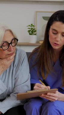 Genç bir kadın doktorun dikey HD videosu, bir kanepede yaşlı bir kadının yanında oturuyor ve tıbbi form doldurmasına yardım ediyor. Sağlık ve Tıp Konsepti 