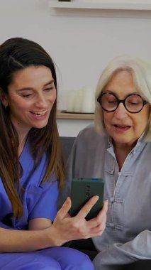 Genç bakıcı hemşirenin akıllı telefon kullanarak ev sağlığı ve teknoloji yardımı sağlayan yaşlı bir kadına yardım ettiği dikey HD videosu.