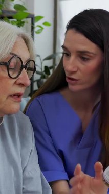 Genç bayan hemşirenin dikey HD videosu, ev ziyareti sırasında evde kanepede oturan yaşlı bir kadına ilaç talimatlarını dikkatlice açıklıyor. Sağlık ve Tıp Konsepti 