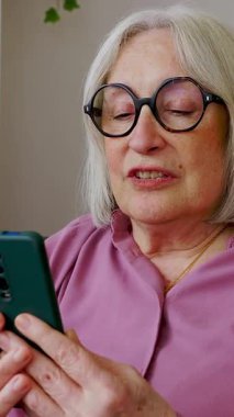 Evde rahat bir koltukta oturmuş modern teknoloji ve iletişimin keyfini çıkarırken, cep telefonu kullanan yaşlı bir kadının dikey HD videosu. Büyükannemin cep telefonundan bir video görüşmesi var.