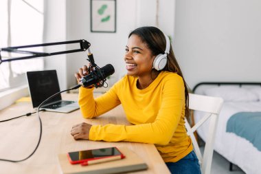 Mutlu genç bir kadın mikrofona konuşuyor, evinin konforundan podcast 'ini kaydediyor, modern teknoloji ve yaratıcılıkla çevrili.