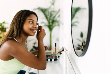 Genç bir kadın aynanın önünde göz farı sürüyor, canlı bir palet ve fırçayla evdeki güzellik rutinini geliştiriyor.