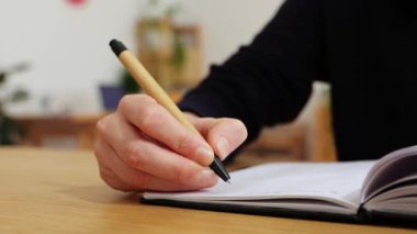 Genç bir kadının, parlak stüdyosundaki ahşap bir masada elinde kalemle deftere el yazısıyla oturduğu yakın plan.