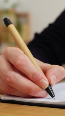 Dikey HD video. Genç bir kadının, parlak stüdyosundaki ahşap bir masada elinde kalemle deftere el yazısıyla oturduğu yakın plan.