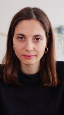 Ev stüdyosundaki masada oturan kameraya gülümseyen genç kadının dikey HD video portresi. Milenyum kadını dişlek bir gülümseme sergiliyor, evde pozitif ve kendinden emin hissediyor.. 