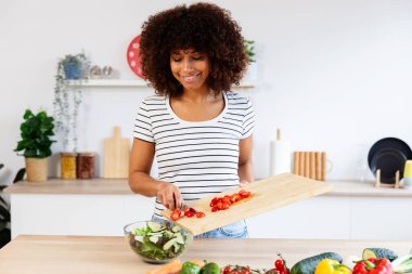 Genç bir kadın modern bir mutfakta sağlıklı bir salata hazırlıyor ve kesme tahtasına taze sebze ekliyor. Sağlıklı yaşam tarzı ve gıda konsepti