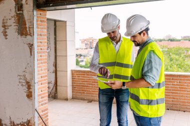 Güvenlik yeleği ve kask giyen iki mühendis inşaat alanında proje detaylarını tartışan dijital tablet kullanıyorlar. Mühendislik ve mimari kavramı