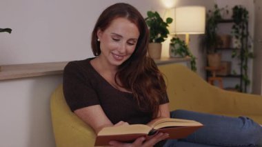 Genç ve güzel bir kadın, modern oturma odasındaki rahat bir kanepede dinlenirken boş zamanlarında kitap okumaktan zevk alıyor.