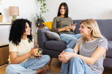 Üç mutlu genç kadın oturma odasında rahatlıyor ve eğleniyor, sağlıklı yemekler yiyor ve sosyal medyada geziniyor. Kadın arkadaşlığı ve gençlik kavramı